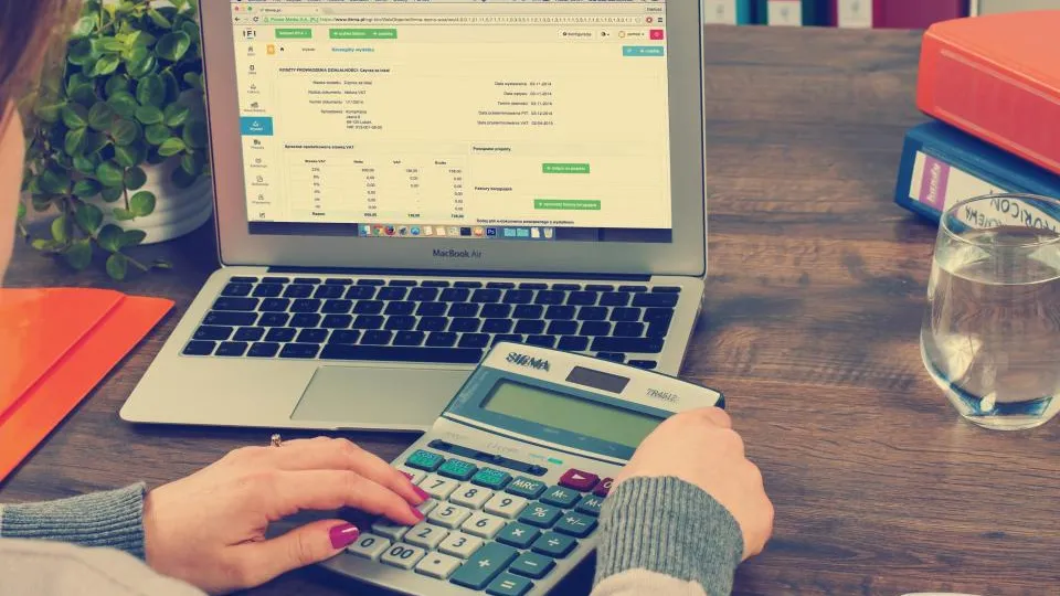Hands using a calculator beside a laptop showing a spreadsheet.